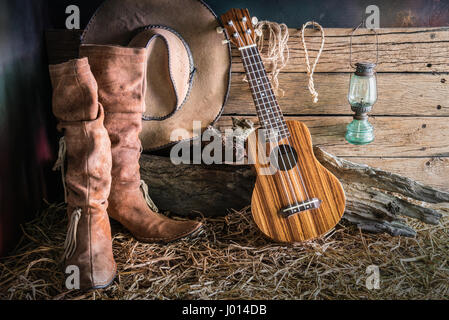 Still life painting photography with ukulele on american west rodeo brown felt cowboy hat and traditional leather boots in vintage ranch barn backgrou Stock Photo