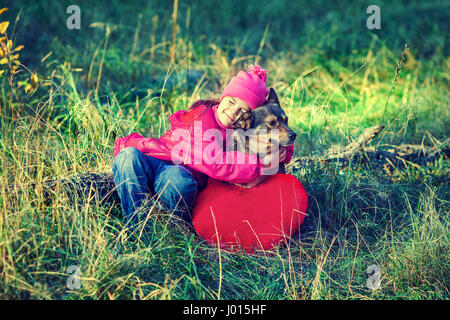 Little girl hugging a dog. Girl sitting on a grass. Near lying red  heart shaped pillow. Conceptual idea. Stock Photo