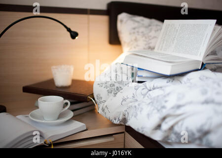 An open book on the bed. Books and coffee Cup on the nightstand. Stock Photo