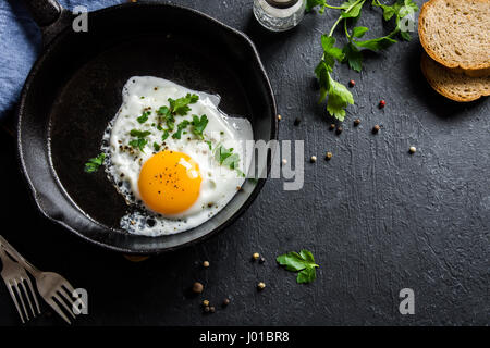 Fried egg. Close up view of the fried egg on a frying pan. Salted and spiced fried egg with parsley on cast iron pan and black background. Stock Photo