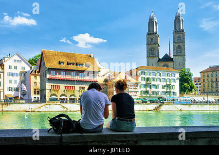 Young couple sitting at Limmatquai and Grossmunster Church in Zurich, Switzerland. Stock Photo