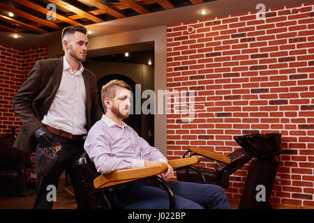 Hairdresser with barber tools standing near a man sitting on chair against brick wall. Stock Photo