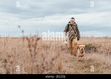 Hiker and small yellow dog walking in grassland on a cloudy day Stock Photo