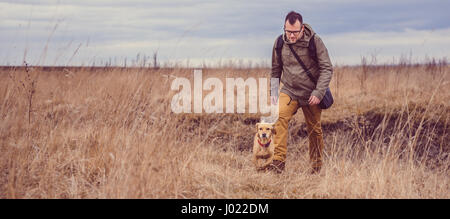 Hiker and small yellow dog walking in grassland on a cloudy day Stock Photo