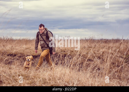 Hiker and small yellow dog walking in grassland on a cloudy day Stock Photo