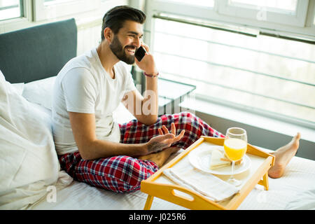 Young man having breakfast in bed and using a mobile phone Stock Photo
