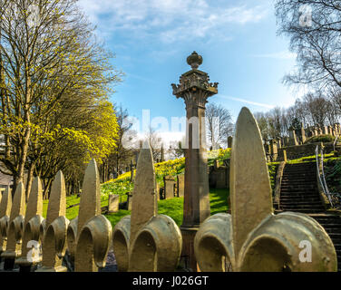 Top of an ornamental cast iron railing with gold fleur de lys making leading line foreground with gravestones and tall monument in Glasgow necropolis Stock Photo