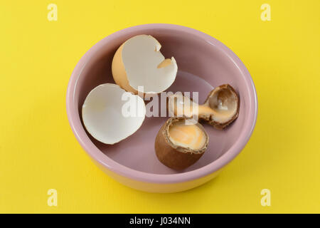 Broken Easter chocolate cream egg with broken eggshells in pink dish against yellow background Stock Photo