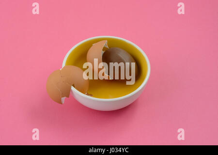Chocolate Easter egg and broken egg shells inside tiny bowl on pink background Stock Photo