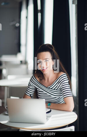 Picture of smiling woman using laptop in a cafe and looking at the camera. Stock Photo