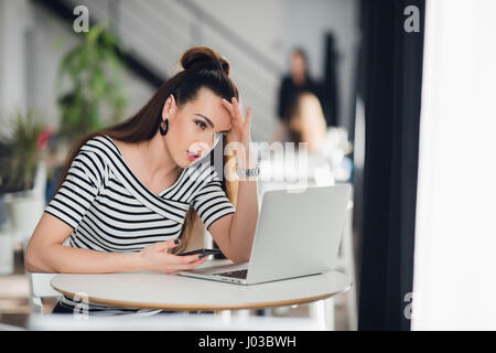 Worried and sad woman got bad news from her relatives in a cafe. Puzzled female looking at a laptop and holding phone. Stock Photo