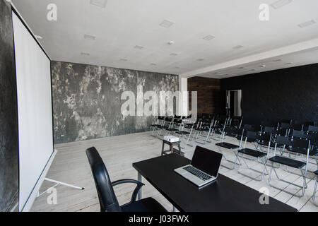 Room for lecture with a lot of dark chairs. Walls are white, loft interior. On the right there is a door. On the background there is a table with a laptop. Stock Photo