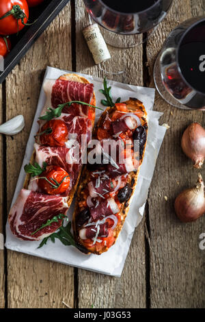 Variety of small sandwiches with jamon, tomatoes, parmesan cheese, fresh basil served with glass of red wine on wooden board, top view. Stock Photo