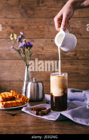 Women's hand with creamer pouring milk in glass with coffee. Cinnamon sticks, homemade cookies and bunch of wildflowers on a wooden background. Breakfast concept. Stock Photo