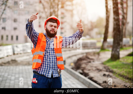 Portrait of brutal beard worker man suit construction worker in safety orange helmet against pavement with showing arms. Stock Photo
