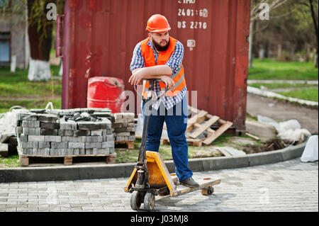 Brutal beard worker man suit construction worker in safety orange helmet with pallet truck. Stock Photo