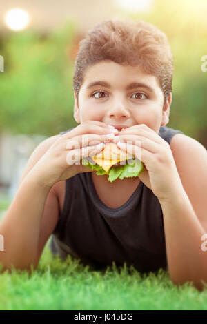 Portrait of a teen boy lying down on fresh green grass in bright sunny day and with pleasure eating tasty burger, happy time in summer camp Stock Photo