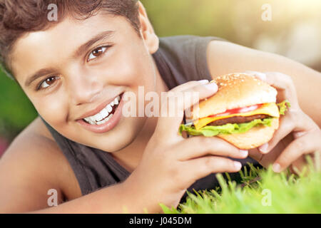 Closeup portrait of a cute happy teen boy lying down on green grass and eating delicious burger, enjoying tasty lunch on sunny day, happy time at summ Stock Photo