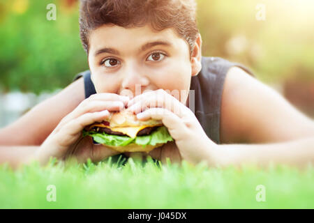 Portrait of a teen boy lying down on the grass on bright sunny day and with pleasure eating tasty burger, happy time in summer camp Stock Photo