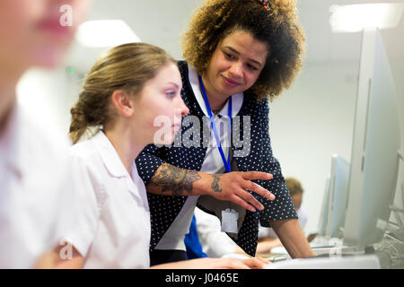 Female teacher with tattoos helping one of her students, who is doing work on a computer. Stock Photo
