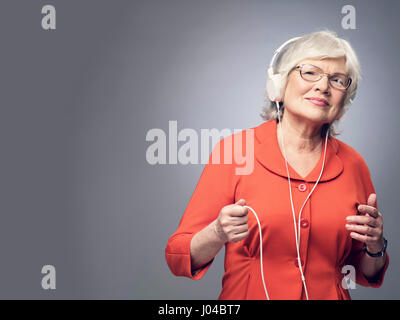 Smiling senior lady with headphones on listening to the music on gray background with copy space Stock Photo