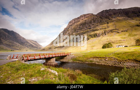 A small bridge crosses the River Coe to Achnambeithach Cottage nestled under the sheer mountains of Glen Coe in the West Highlands of Scotland.
