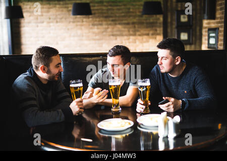 Three young men in casual clothes are talking, eating chips and drinking beer while sitting in pub, close-up Stock Photo