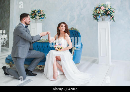 Bride in beautiful dress and groom in gray suit sitting on sofa indoors in white studio interior like at home. Trendy wedding style Stock Photo