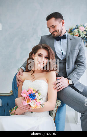 Bride in beautiful dress and groom in gray suit sitting on sofa indoors in white studio interior like at home. Trendy wedding style Stock Photo