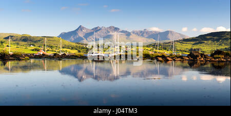 Boats shelter in Portree Bay on Scotland's Isle of Skye, with the Cuillin mountains providing the backdrop, reflected in the blue waters. Stock Photo