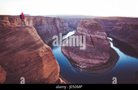 A male hiker is standing on steep cliffs enjoying the beautiful view of Colorado river flowing at famous Horseshoe Bend overlook in twilight, Arizona Stock Photo