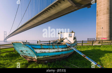 A derelict wooden boat under the Humber Bridge which spans the Humber estuary and viewed on a spring morning in Hessle, near Hull Stock Photo