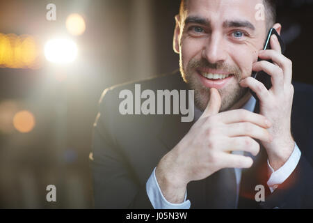 Portrait of modern handsome businessman using smartphone making call and smiling at camera while talking against black background  with lens flare Stock Photo