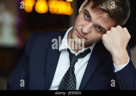 Portrait of young tired businessman wearing formal suit looking at camera with serious neutral expression resting head on hand Stock Photo