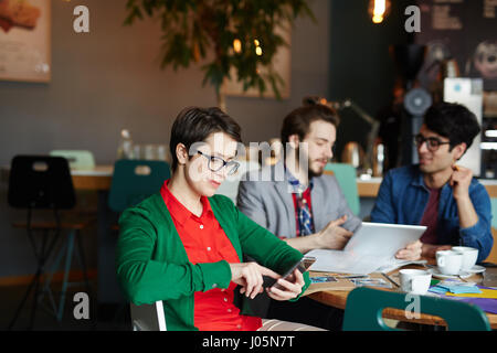 Portrait of creative young woman wearing glasses using smartphone looking at screen at table in cafe with colleagues Stock Photo