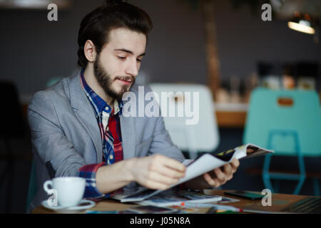 Portrait of confident long haired man wearing business casual clothes sitting at table in cafe reading magazines Stock Photo