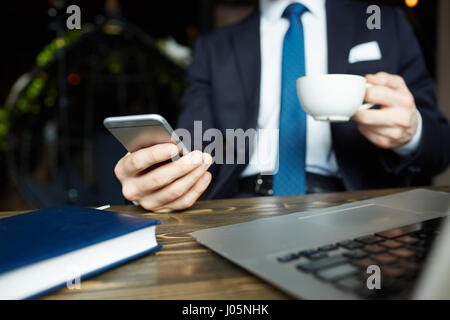 Closeup low angle of unrecognizable elegant businessman working in cafe, holding mobile phone, texting and drinking coffee from cup Stock Photo