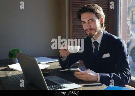 Portrait of young handsome businessman working with smartphone and laptop at wooden sunlit table during coffee break, looking at camera and smiling ch Stock Photo