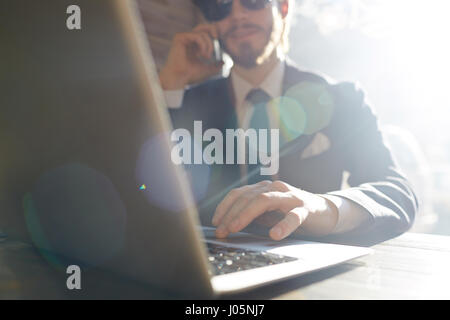 Portrait of cool businessman working using smartphone and laptop at sunlit table in cafe, wearing big sunglasses and drinking coffee in bright sunligh Stock Photo