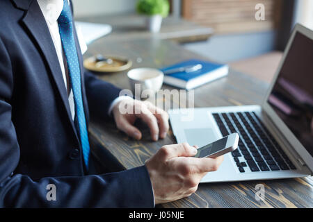 Side view closeup of elegant modern businessman during coffee break: holding  smartphone and typing text messages while working with laptop in cafe Stock Photo