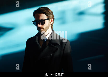 Portrait of handsome man wearing sunglasses and black coat in bright sunlight against blue background, looking away from camera Stock Photo