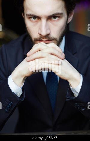 Head and shoulders portrait of handsome bearded man wearing elegant business suit, looking strongly at camera resting chin on clasped hands Stock Photo