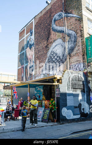 Ethiopian vegan cuisine stalls on Hanbury Street just off Brick Lane in East London. Above is a large street art painting of a heron. Stock Photo