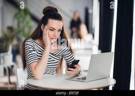 Worried and sad woman got bad news from her relatives in a cafe. Puzzled female looking at a laptop and holding phone. Stock Photo