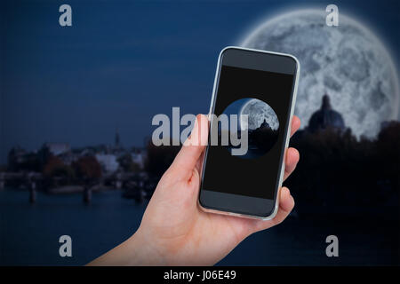 Hand holding mobile phone against white background against large moon over river city Stock Photo