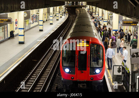 A train arrives at the platform of Aldgate East London Underground Station where passengers are waiting. Stock Photo