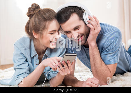 Smiling young couple lying on floor and using smartphone with white headphones Stock Photo