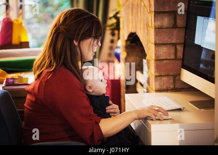 A woman seated with a baby on her lap using her computer, both watching the screen. Stock Photo