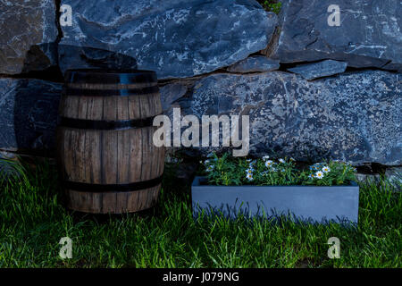 Night shot of a vintage oak barrel on the lawn beside a pot of daisy flowers with natural stone wall as background Stock Photo
