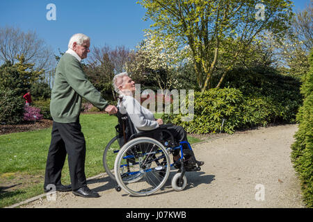 Retired husband taking disabled elderly wife in wheelchair for a walk in the park on a sunny day in spring Stock Photo